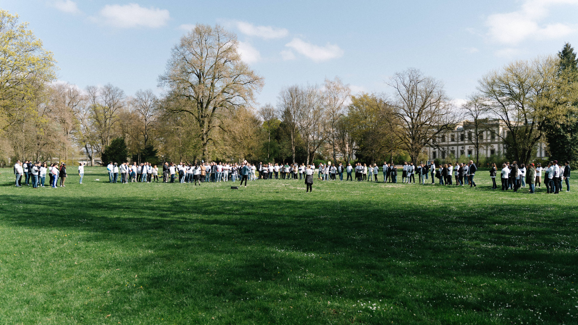 A large group standing on green gras, while getting informations about an event.
