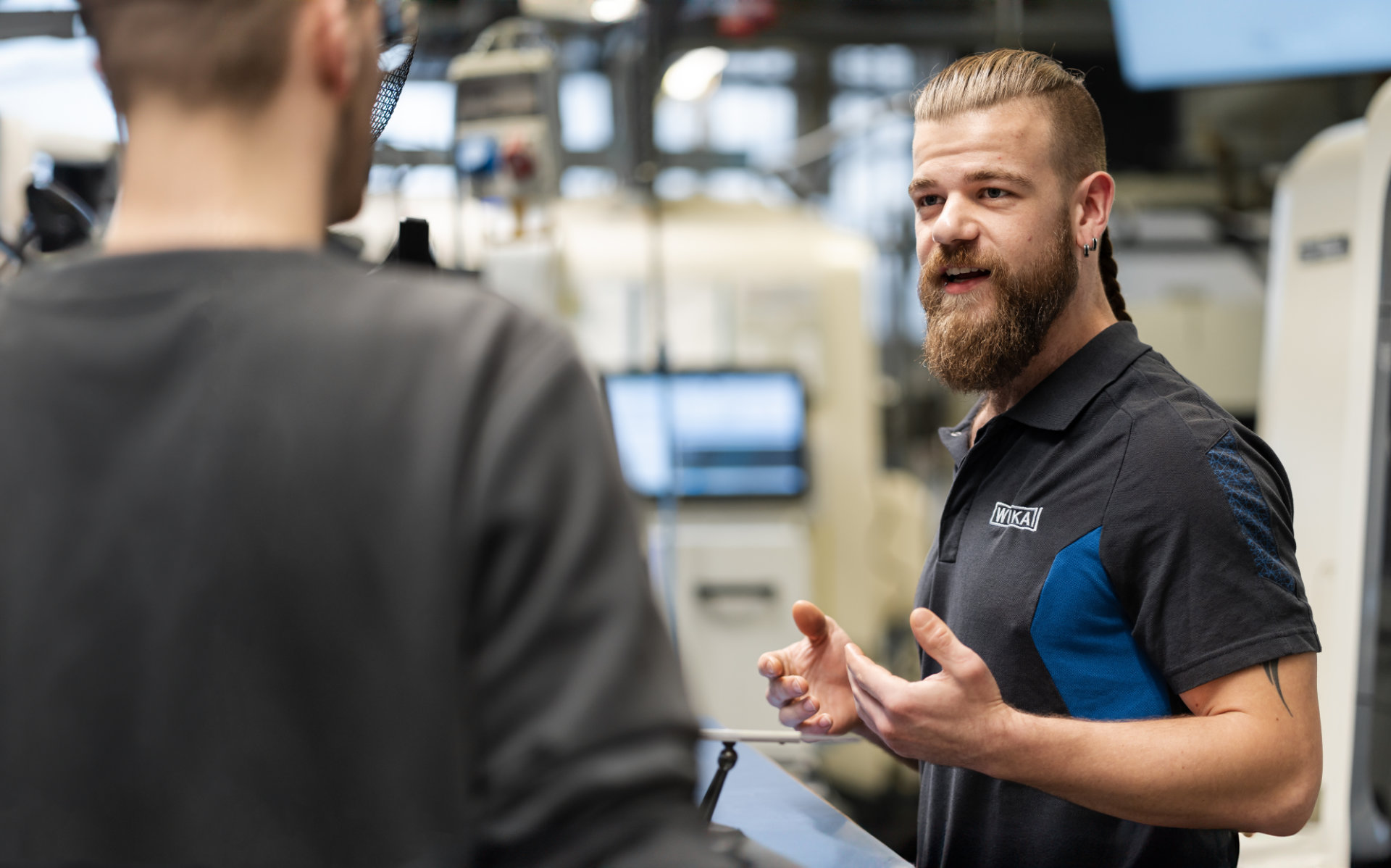 A bearded technician explaining something to a colleague at work with industrial machinery.