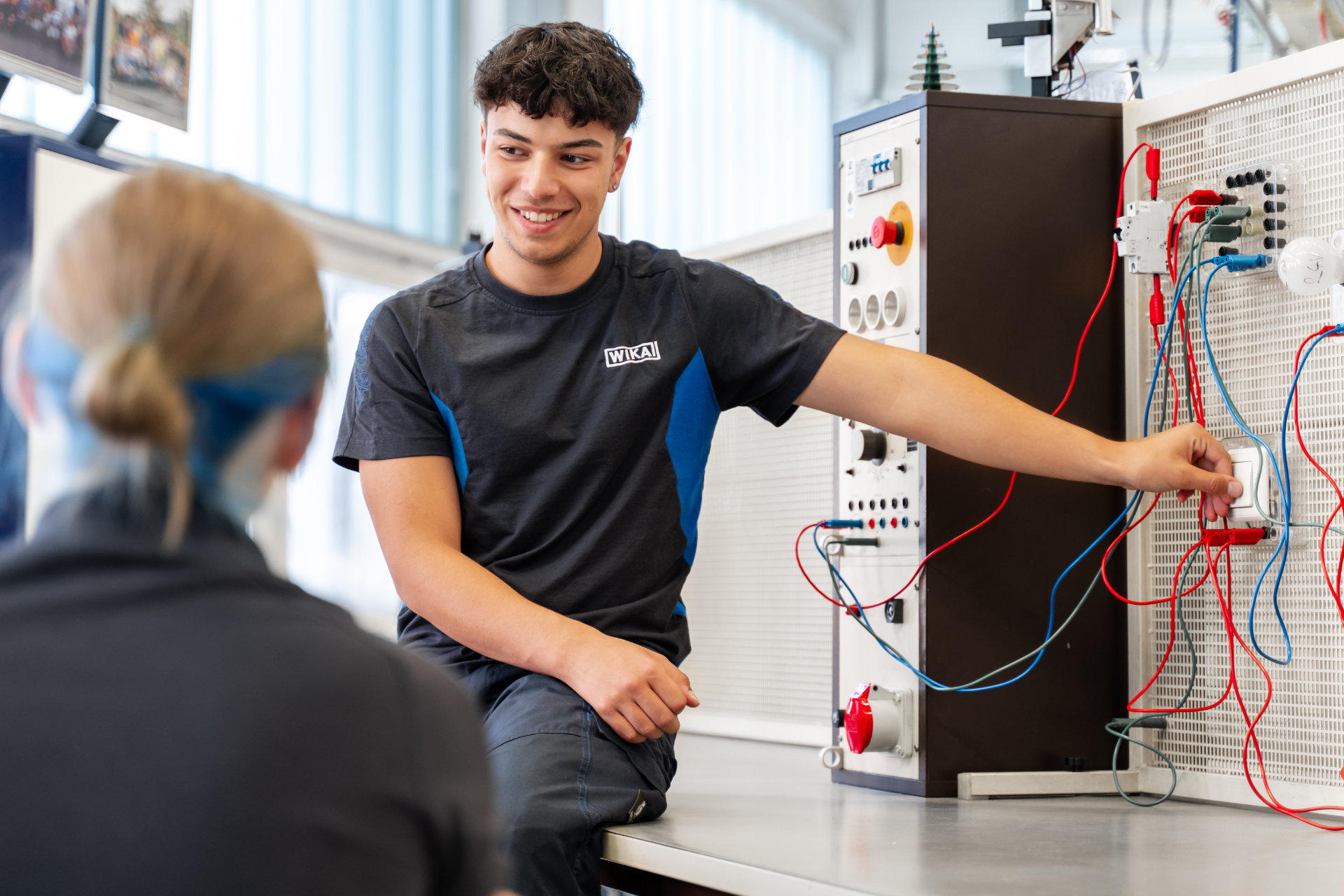 A young man in a dark t-shirt with a logo smiles while pointing to something on an electrical training module during a lesson, with an attentive student or colleague in the foreground, facing the person.