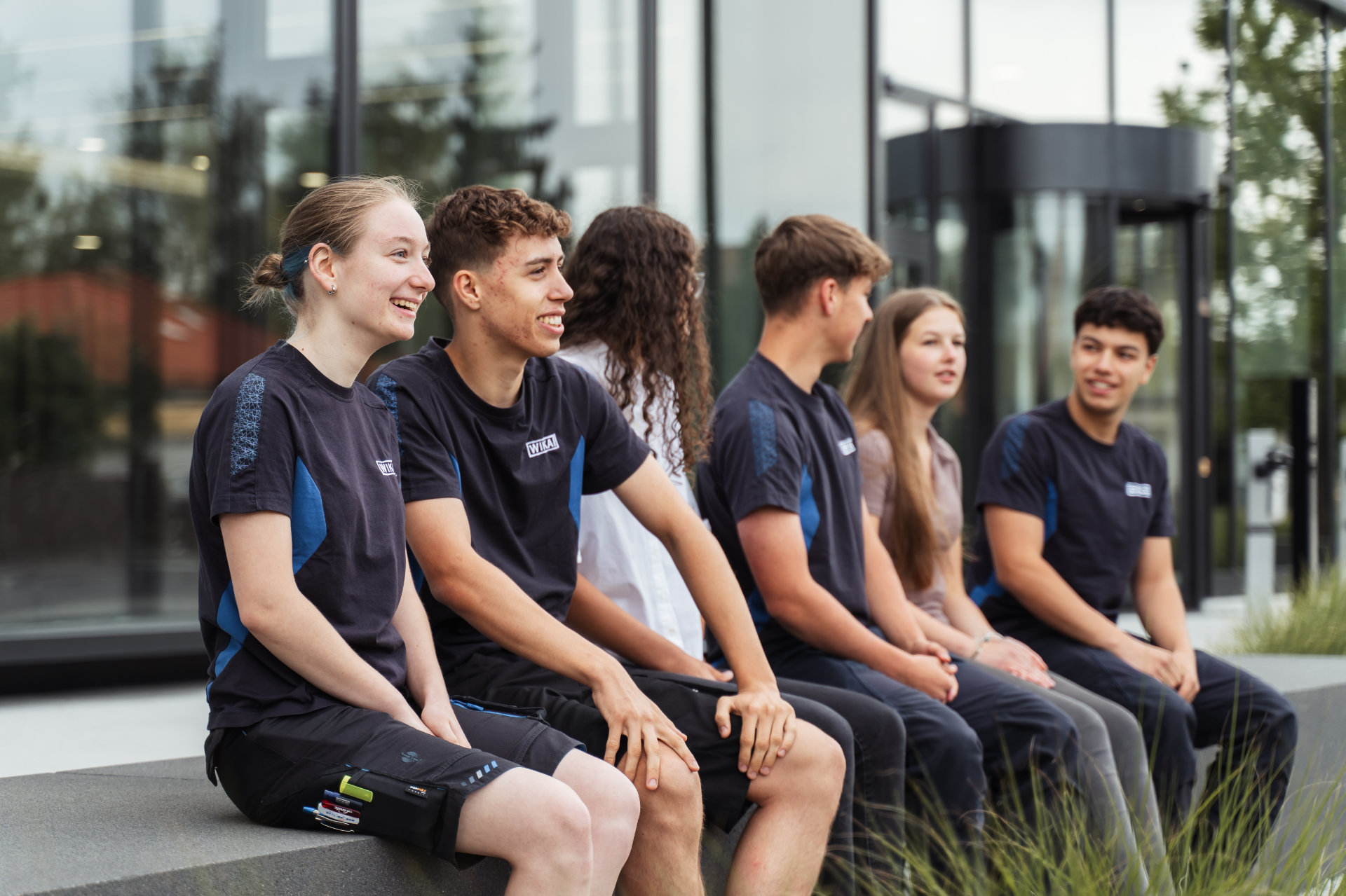 A group of smiling young adults, wearing matching sporty attire sitting in a row outdoors, seemingly enjoying a light-hearted conversation or shared moment.