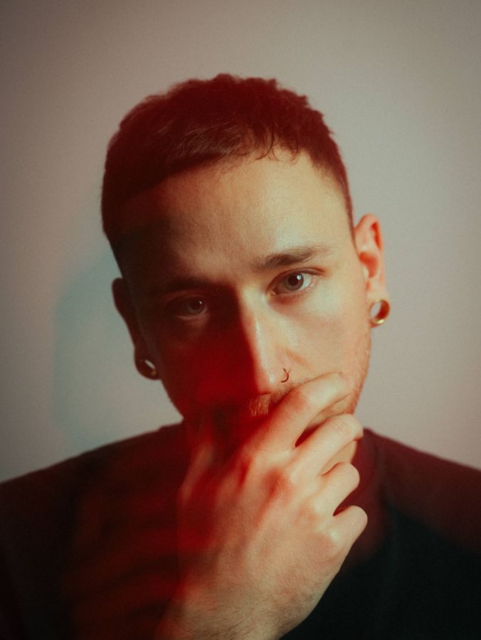 A photographer with short hair and hoop earrings gazes thoughtfully, partially covering their mouth with one hand. The lighting casts a soft red glow on their face, creating a warm and contemplative atmosphere that highlights the art of videography.