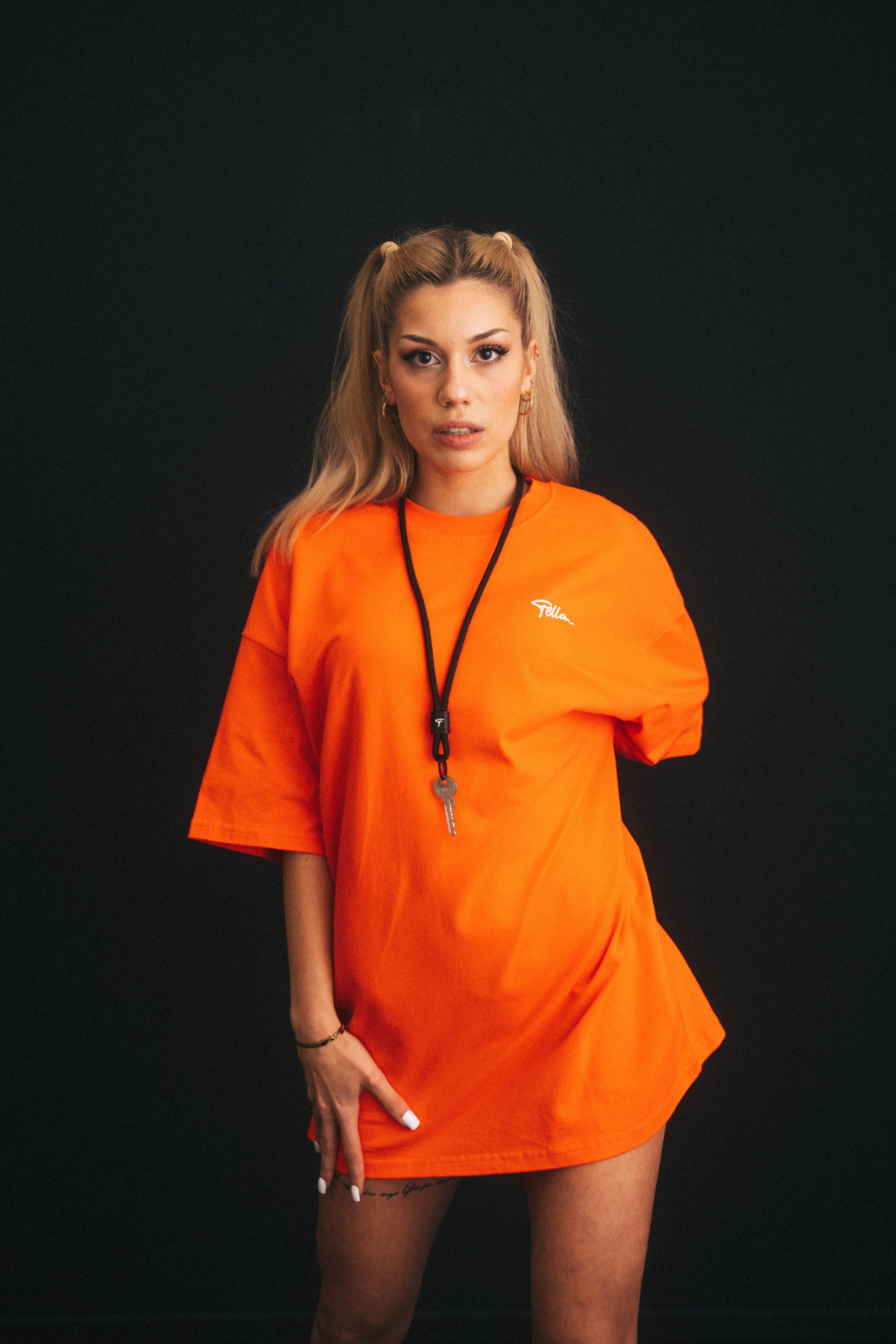 A self-confident young woman in a bright orange, oversized T-shirt stands in front of a dark background, captured by an event photographer from Aschaffenburg, her gaze directed at the camera with subtle intensity.