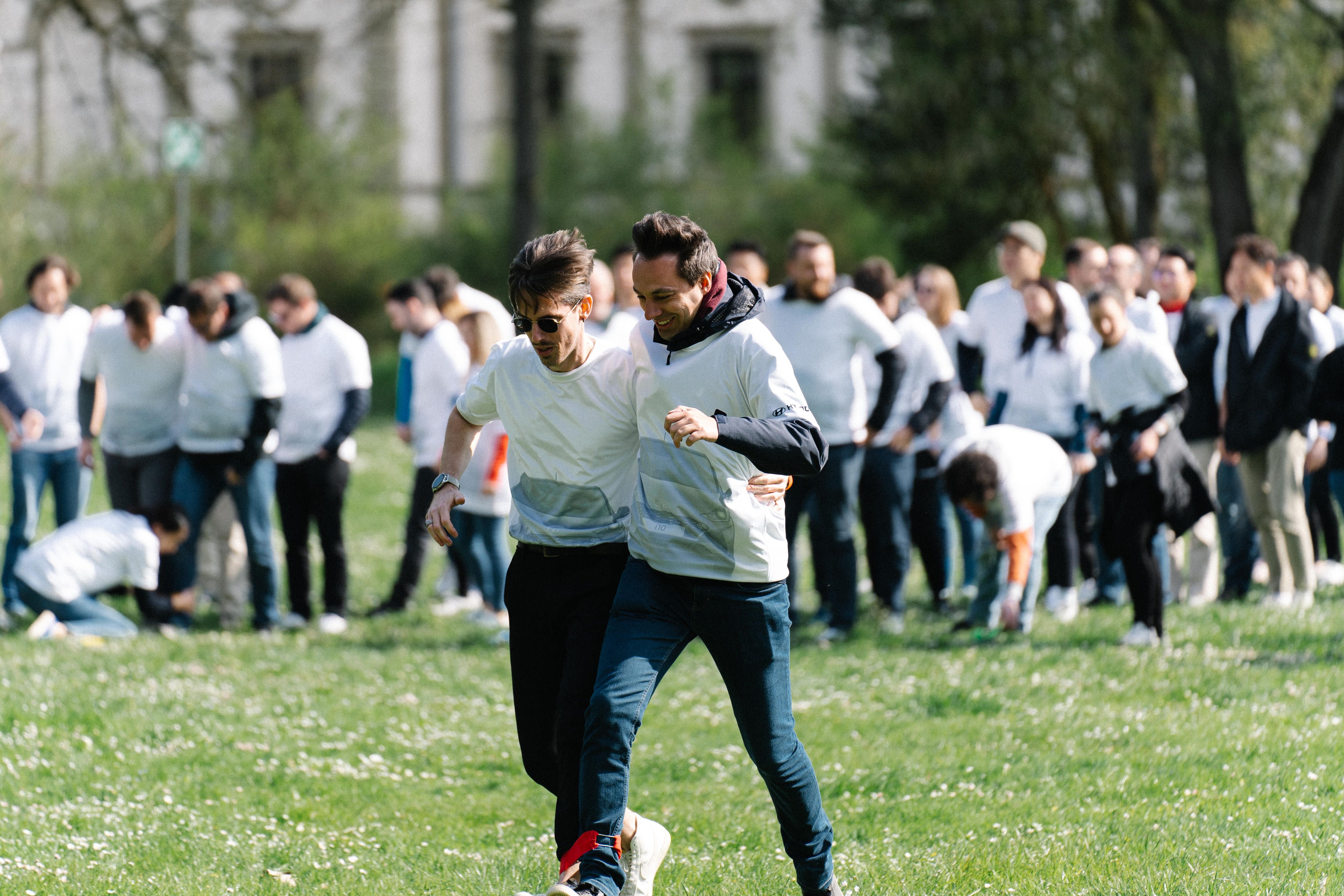 Two individuals participate in a three-legged race, tightly bound together at the leg, striving for coordination and balance, as spectators in the background watch their effortful sync. 