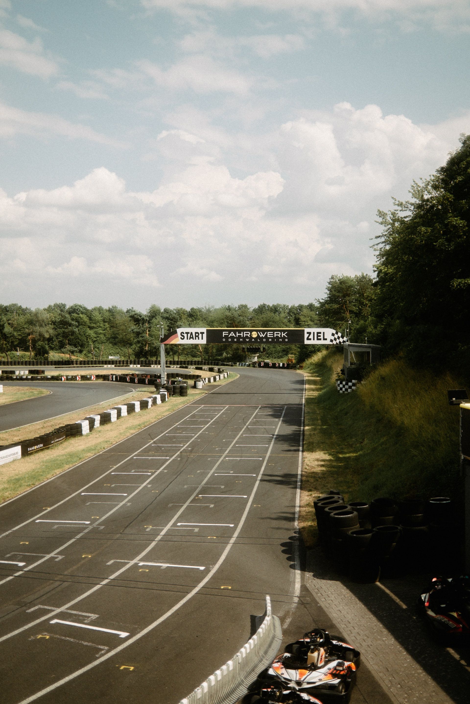 A serene racetrack awaits the thrill of speed, flanked by lush greenery on a sunny day, as go-karts prepare to zip across the start-finish line, captured through the lens
