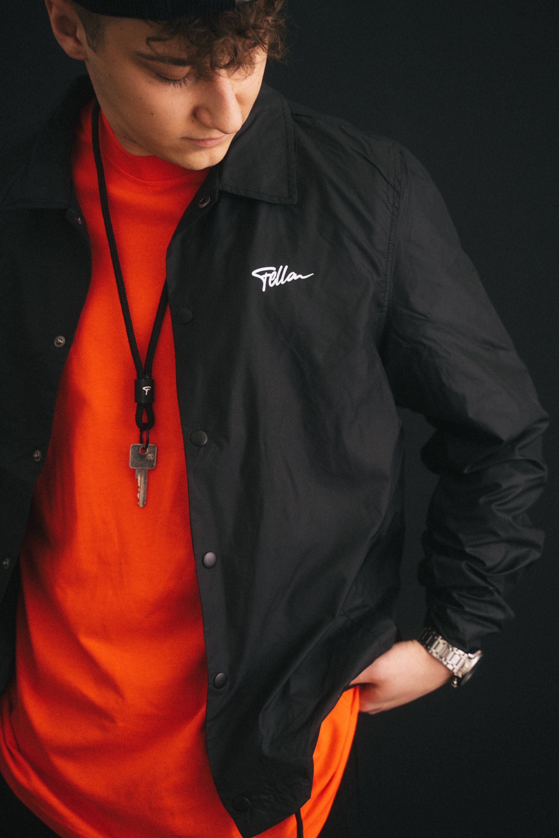 Young man in a stylish black jacket and bright orange shirt, looking down thoughtfully, with a necklace and a watch, photographed by an experienced photographer from Aschaffenburg.