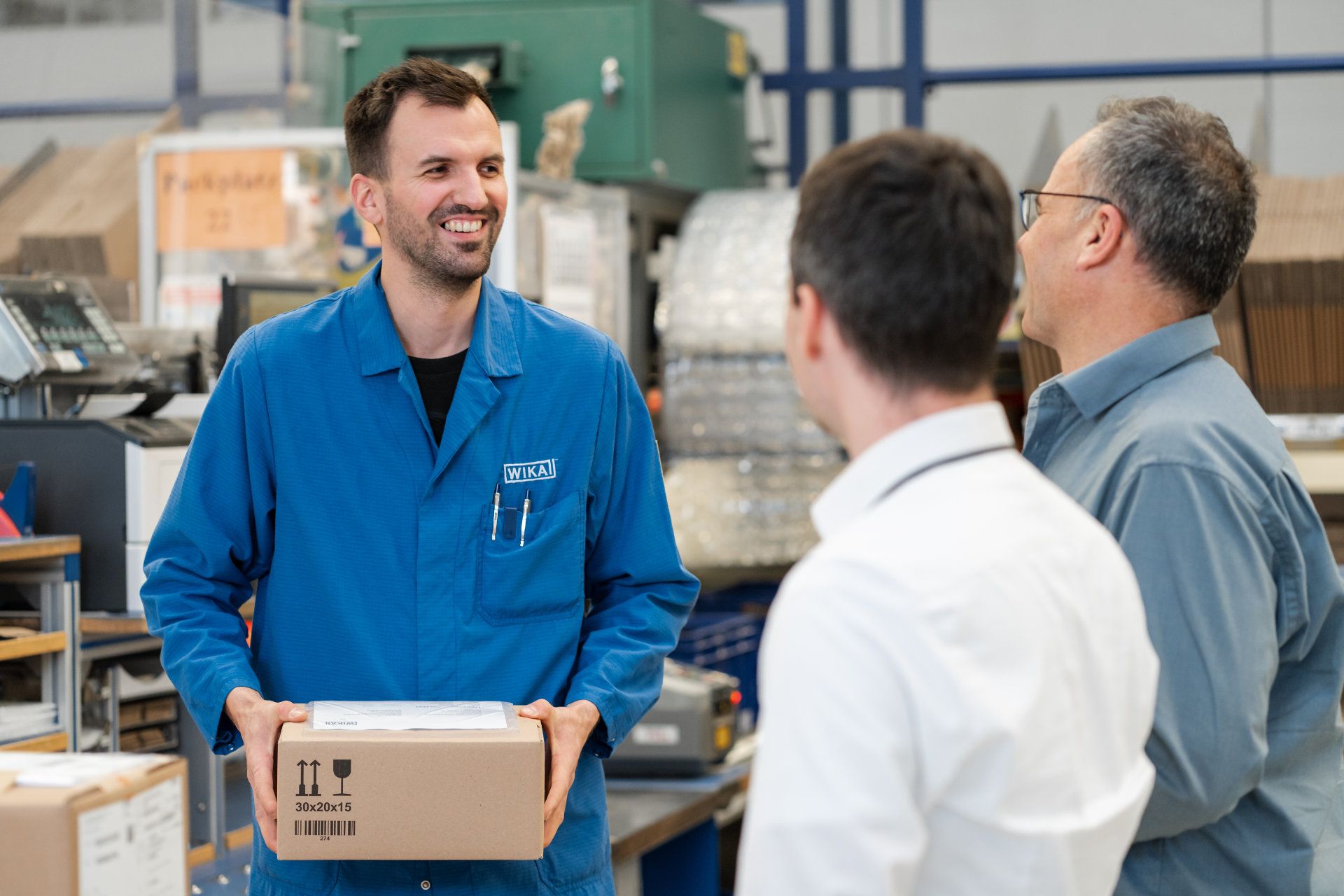 Three colleagues sharing a light-hearted moment in a warehouse, with one person in a blue work shirt holding a package and engaging in a conversation with the others.