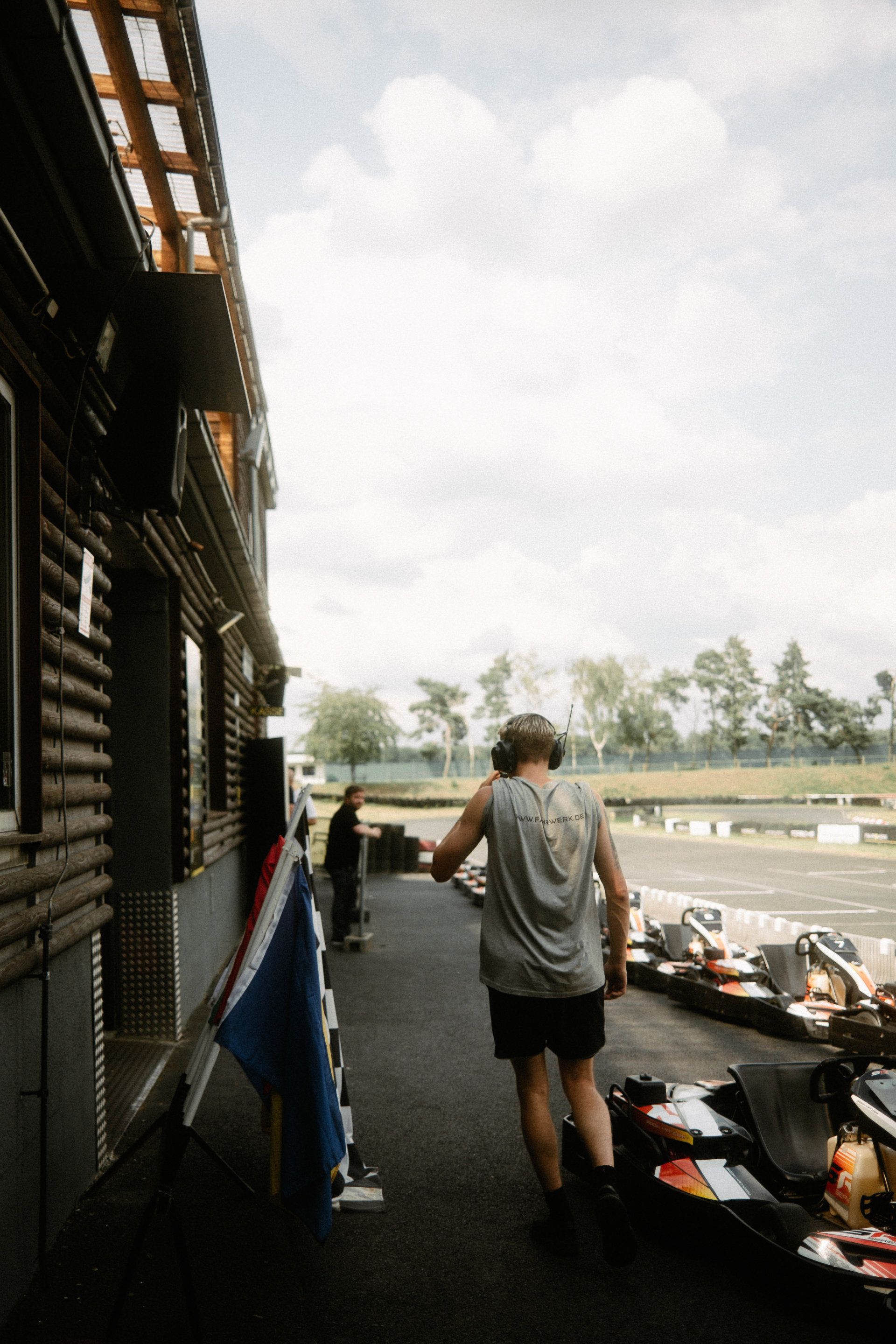 A person strolling down the pit lane at a go-kart track, with go-karts lined up ready for action, while a videographer captures every dynamic moment.