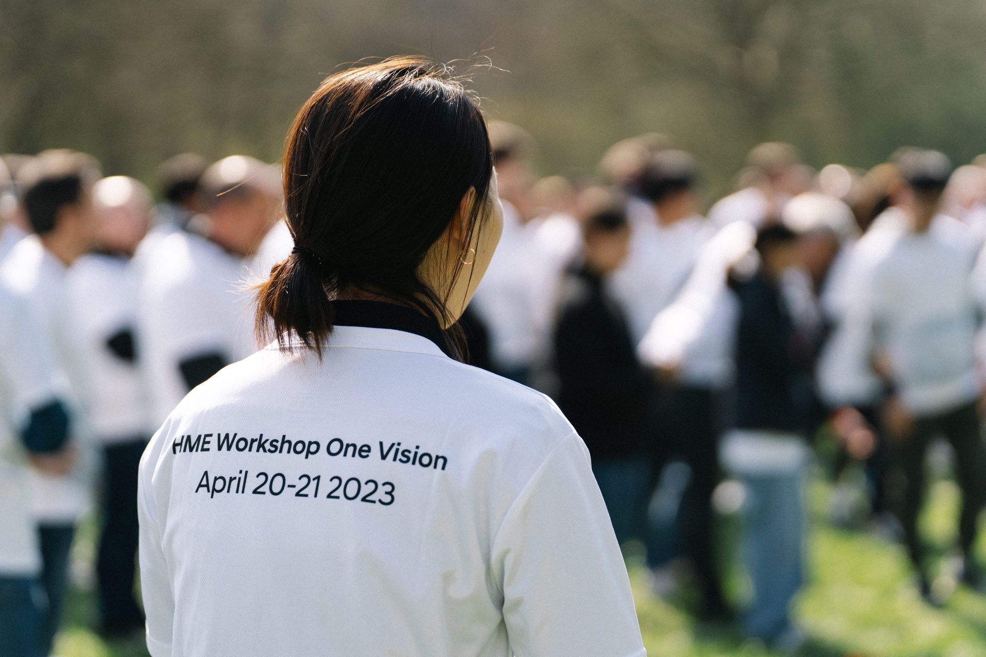 A photographer from Aschaffenburg captures a woman wearing a shirt labeled "hme workshop one vision april 20-21 2023" as she stands with her back to the camera, facing a group of people.