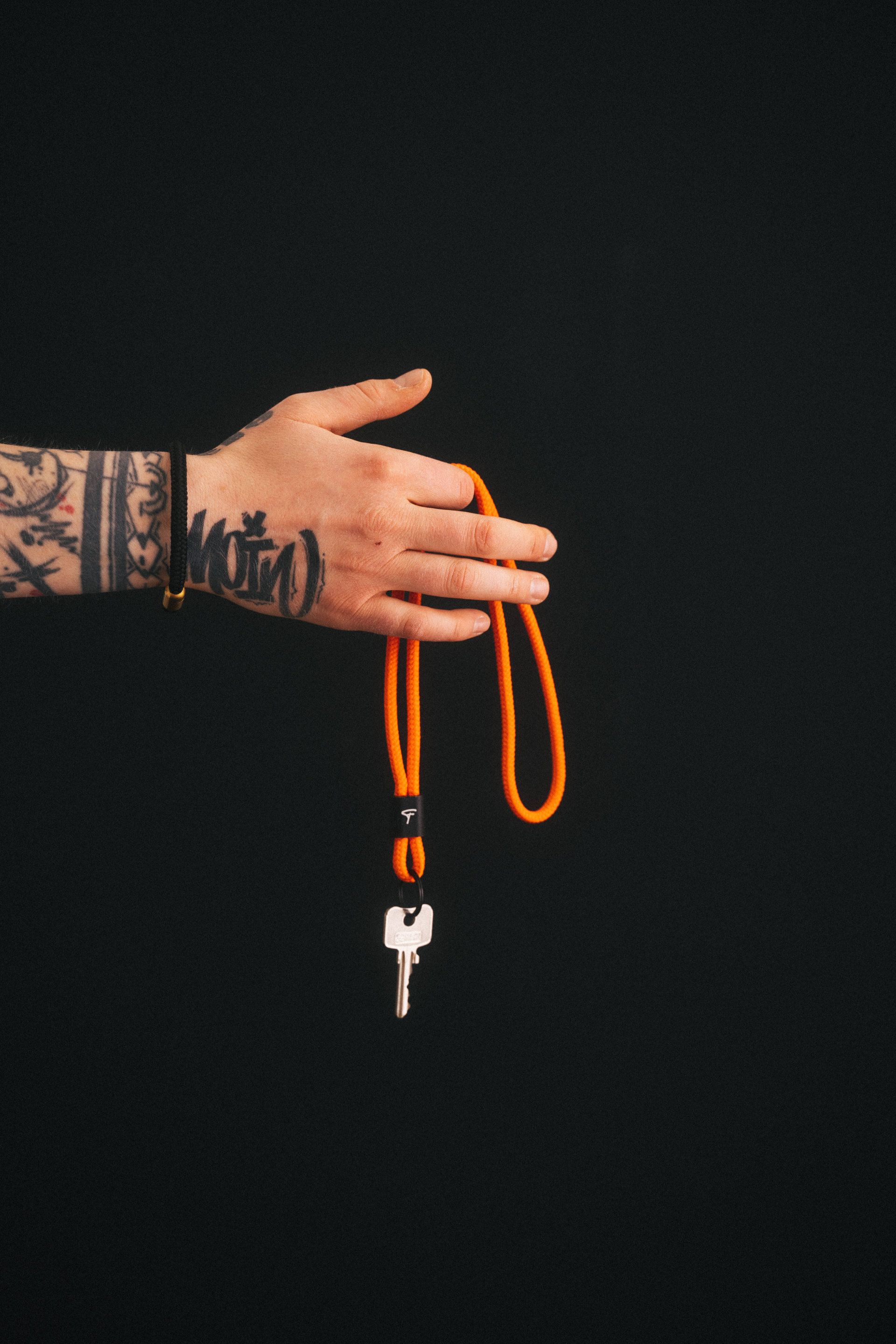 A tattooed hand holding an orange-coloured lanyard with a loop and a single key on a black background.