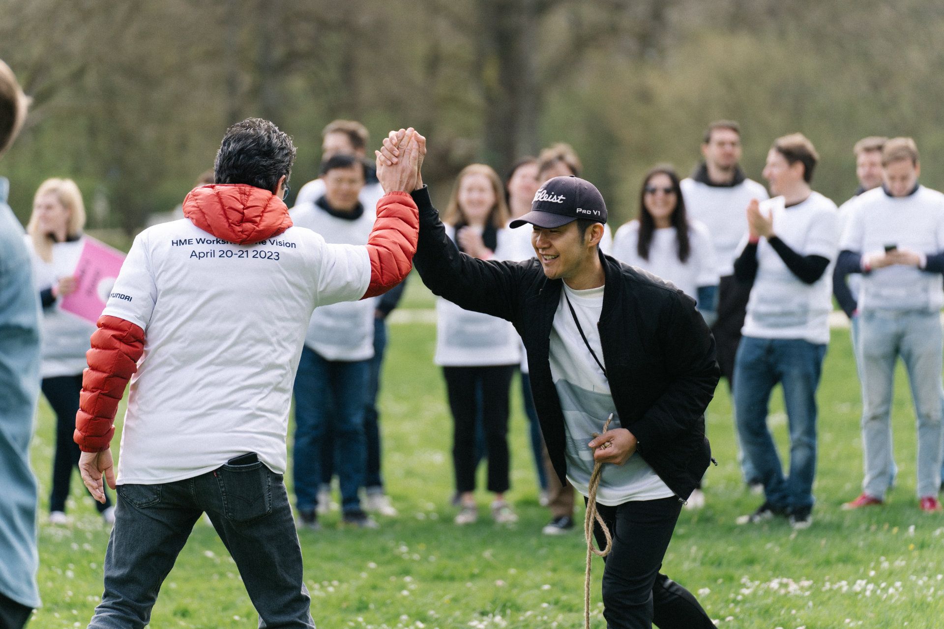 Two individuals exchanging a high-five in a park while surrounded by a group of onlookers wearing coordinated t-shirts, suggesting a team-building activity.