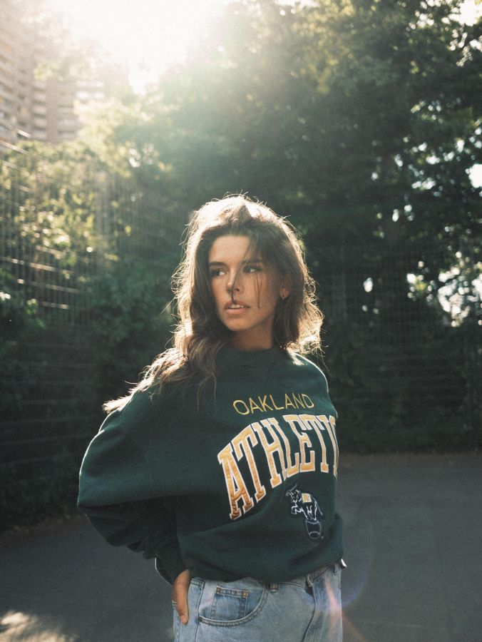 A young woman bathed in soft sunlight stands pensively in an urban park setting, clad in a casual Oakland Athletics sweatshirt and jeans.